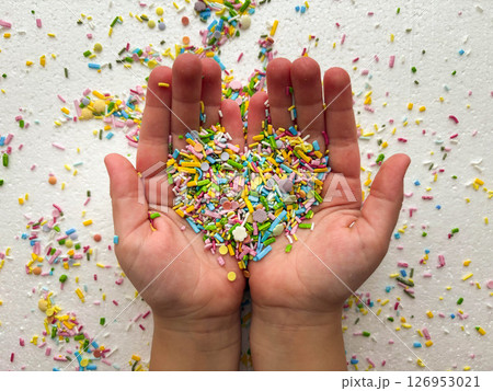 Child's hands are covered with multi-colored sugar sprinkles and form a heart shape. Child plays with small sprinkles topping. Child's palms on a white background. Child's hands are covered with multi-colored sugar sprinkles and form a heart shape. Child plays with small sprinkles topping. Child's palms on a white background. 126953021