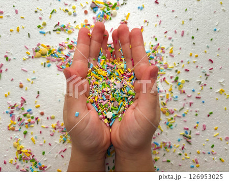 Child playing with colored sugar candies on white background. Confectionery sprinkles in child's palms. Children's game, fine motor skills, mess. 126953025