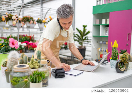 A Caucasian man in an apron works at the counter of a flower shop, using a laptop and taking notes while on the phone. A Caucasian man in an apron works at the counter of a flower shop, using a laptop and taking notes while on the phone. 126953902