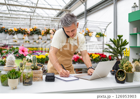 A Caucasian man working at a florist shop is on the phone while taking notes and using a laptop on a bright day. 126953903