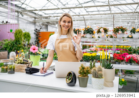 A smiling florist gestures with an 'OK' sign while standing at the sales counter, in her shop surrounded by plants and flowers. A smiling florist gestures with an 'OK' sign while standing at the sales counter, in her shop surrounded by plants and flowers. 126953941