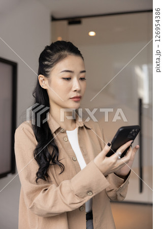 Serious Vietnamese professional woman standing in workplace with smartphone Serious Vietnamese professional woman standing in workplace with smartphone 126954386