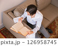 young dandy man in a white shirt reading a book while seated on a beige couch in a cozy living room setting during daytime. top view 126958745
