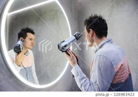 A man with damp hair styles himself using a hairdryer while looking into a round LED-lit mirror in a clean, modern bathroom interior. 126963422