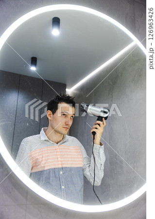 Young man stands in front of a glowing round mirror, carefully drying and styling his wet hair with a hairdryer in a modern bathroom. 126963426