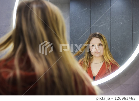 Young woman in a red robe pauses while holding a toothbrush, staring at her reflection in a circular LED mirror inside a sleek modern bathroom. 126963465