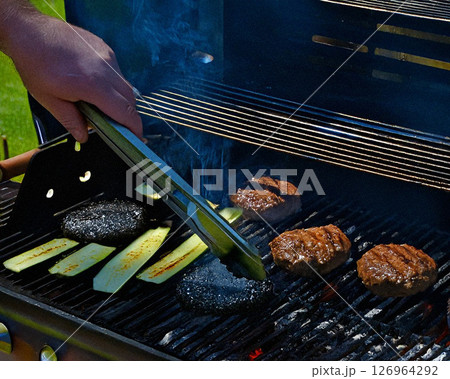 A hand uses tongs to turn vibrant yellow bell peppers on a sizzling gas grill, surrounded by well-cooked burgers, zucchini, and tomatoes. 126964292