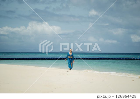 Girl in blue beach dress walking on white beach against blue ocean and sky 126964429