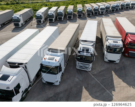 Aerial view of a large truck parking lot at a logistics hub, with multiple commercial semi trucks parked in organized rows. Aerial view of a large truck parking lot at a logistics hub, with multiple commercial semi trucks parked in organized rows. 126964625