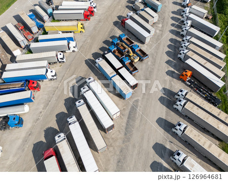 Aerial view of a large truck parking lot at a logistics hub, with multiple commercial semi trucks parked in organized rows. Aerial view of a large truck parking lot at a logistics hub, with multiple commercial semi trucks parked in organized rows. 126964681