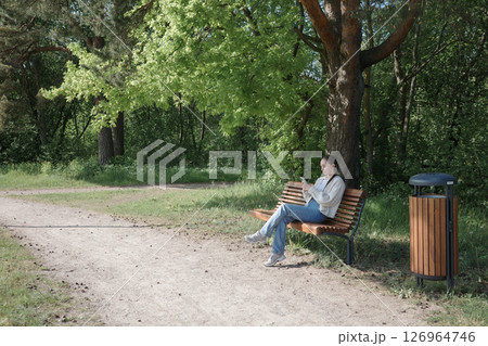Young woman sitting on a bench in a sunny park using a smartphone. Concept of digital communication, leisure, outdoor lifestyle, springtime relaxation and solo travel. 126964746