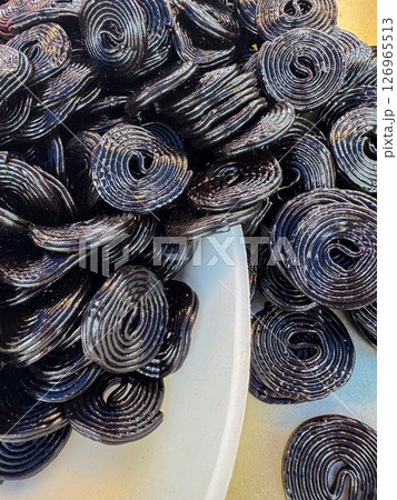 Black licorice wheels are stacked in a white bowl, highlighting their unique swirl pattern and glossy surface. The close-up view emphasizes the texture and color contrast 126965513