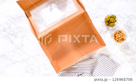 Flat lay. Hands are meticulously adding fresh red grapes to a bowl, complementing a beautifully arranged charcuterie box brimming with a variety of cheeses, olives, and cured meats, set against a 126968706