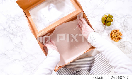 Flat lay. Hands are meticulously adding fresh red grapes to a bowl, complementing a beautifully arranged charcuterie box brimming with a variety of cheeses, olives, and cured meats, set against a Flat lay. Hands are meticulously adding fresh red grapes to a bowl, complementing a beautifully arranged charcuterie box brimming with a variety of cheeses, olives, and cured meats, set against a 126968707