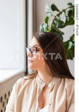 body positive woman psychologist with long hair and glasses is seated by a window, gazing thoughtfully into the distance. 126968941