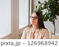 Woman in glasses sits by the window, enjoying quiet reflection and natural light in a modern indoor setting with plants. body positive woman psychologist 126968942