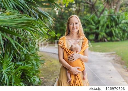Mother holding her little baby in a yellow sling in the park. Warm and loving family moment. Babywearing, parenting, and mother-child bonding concept Mother holding her little baby in a yellow sling in the park. Warm and loving family moment. Babywearing, parenting, and mother-child bonding concept 126970798