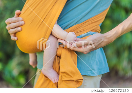 Father holding his little baby in a yellow sling in the park. Warm and loving family moment. Babywearing, fatherhood, and parent-child bonding concept 126972120