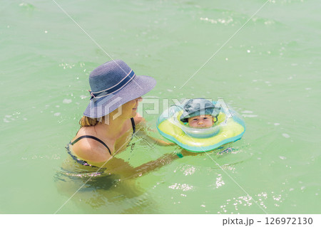 Baby floating in a neck ring in turquoise water with mom nearby. Early swimming and water safety concept 126972130