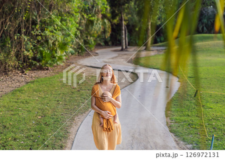 Mother holding her little baby in a yellow sling in the park. Warm and loving family moment. Babywearing, parenting, and mother-child bonding concept 126972131