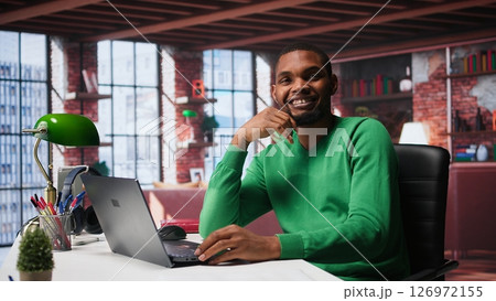 African american remote worker solving his daily freelancing tasks on laptop, working in relaxation from the comfort of his modern loft. Tech savvy black man internet user, multitasking. Camera B. 126972155