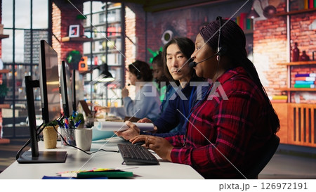 Experienced call center team leader assisting employee and drinking coffee in office. Asian account manager doing teamwork with colleague, enjoying beverage in workplace, camera B Experienced call center team leader assisting employee and drinking coffee in office. Asian account manager doing teamwork with colleague, enjoying beverage in workplace, camera B 126972191