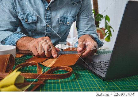Craftsman measuring and cutting leather in a workshop with a laptop 126973474