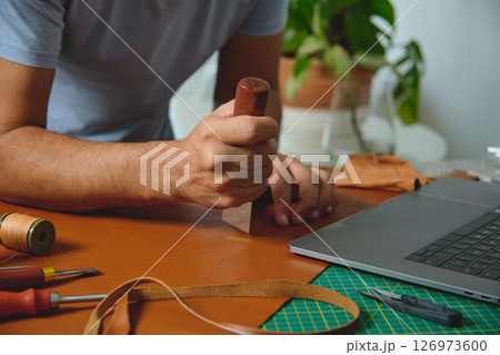 Shoemaker working leather with craft tools and laptop in his workshop 126973600