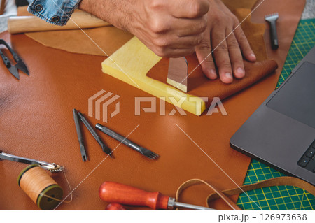 Shoemaker crafting leather using traditional tools and laptop in workshop 126973638