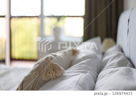 Cozy bedroom interior, beige bedspread and pillow, bedside lamp next to the bed, light from the window 126974415