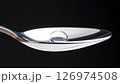 A macro shot of a single drop of liquid or a clear pill on a silver spoon against a black background. 126974508