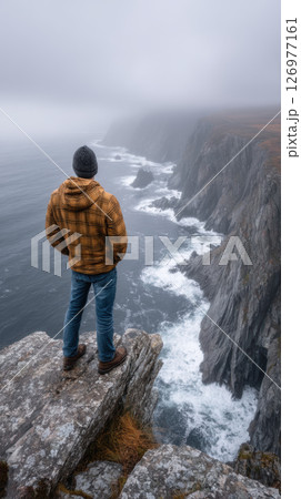 Man standing on cliff edge overlooking dramatic ocean coastline, misty weather, adventure mood Man standing on cliff edge overlooking dramatic ocean coastline, misty weather, adventure mood 126977161
