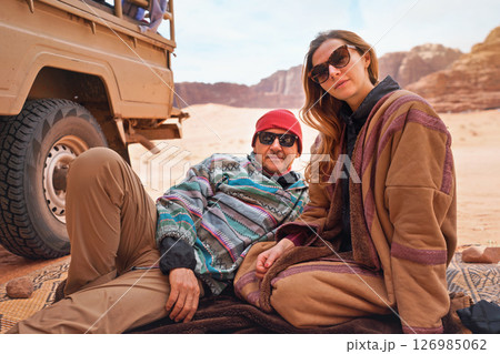 Man and younger woman wearing traditional Bedouin warm coat - bisht -  sitting or laying on ground blanket near off road vehicle back, blurred Wadi Rum desert background 126985062