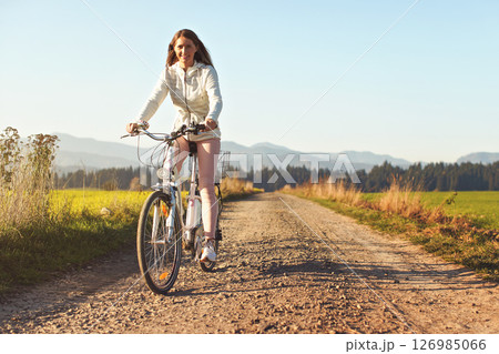 Young woman rides bicycle on dusty road towards camera, afternoon sun shines to fields and forest in background 126985066