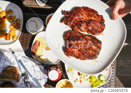 Two freshly grilled pork loin steaks on white plate held by hand over garden table, with more food from grill. Two freshly grilled pork loin steaks on white plate held by hand over garden table, with more food from grill. 126985078