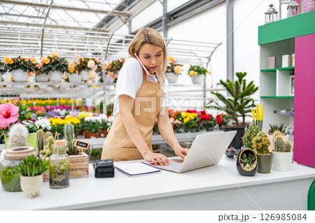 A Caucasian woman works on a laptop while on the phone at a flower shop. She wears an apron and is surrounded by plants and flowers. 126985084