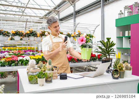 A Caucasian florist at his shop, taking a photo of a vibrant hibiscus flower for sale. Various plants are arranged on display. 126985104