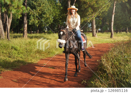 Young woman in shirt and straw hat, riding small black horse in the park, blurred background with houses and trees 126985116