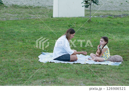 A woman and a young girl sit on a blanket in a grassy field, seemingly engaged in quiet play or conversation. 126986521