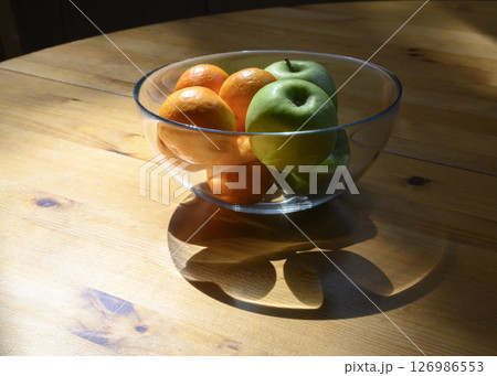 Green apples and orange tangerines in glass vase casting shadow in rays of the sun on wooden table 126986553