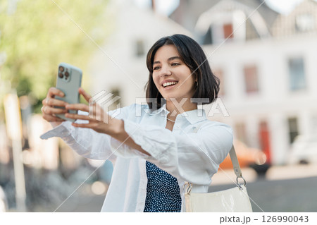 Snap moment. Multiethnic woman with a bright smile and short hair takes a selfie in a white shirt on a lively street with modern architecture 126990043