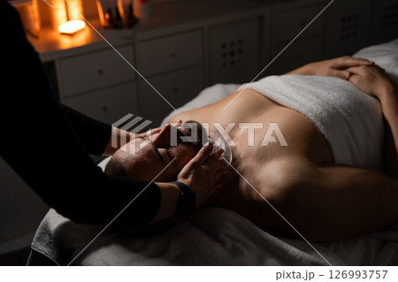 A man is lying on a massage table covered with a towel while receiving a facial spa treatment. A professional is gently applying a towel or compress to his face. The atmosphere is calm and relaxing 126993757