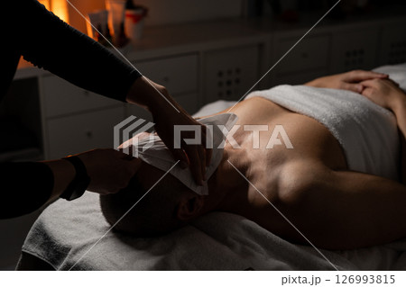 A man is lying on a massage table covered with a towel while receiving a facial spa treatment. A professional is gently applying a towel or compress to his face. The atmosphere is calm and relaxing A man is lying on a massage table covered with a towel while receiving a facial spa treatment. A professional is gently applying a towel or compress to his face. The atmosphere is calm and relaxing 126993815