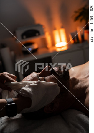 A man is lying on a massage table covered with a towel while receiving a facial spa treatment. A professional is gently applying a towel or compress to his face. The atmosphere is calm and relaxing 126993820