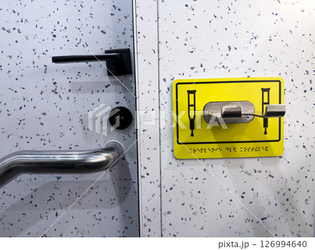 Stainless steel crutch holder with tactile braille sign mounted on a speckled restroom wall near a support handle and door. Designed for accessibility and inclusive infrastructure in public spaces Stainless steel crutch holder with tactile braille sign mounted on a speckled restroom wall near a support handle and door. Designed for accessibility and inclusive infrastructure in public spaces 126994640