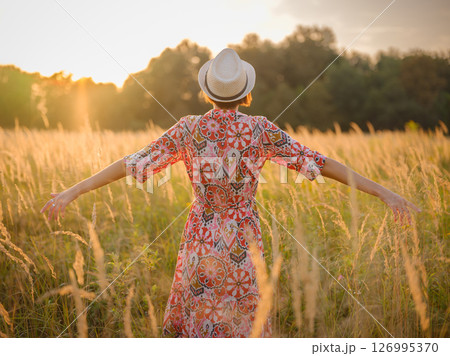 Young woman walking through picturesque European field in late summer. Golden sunlight, lush greenery, and serene rural atmosphere create peaceful countryside scene. 126995370