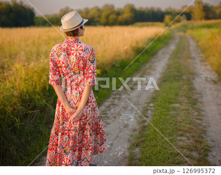 Young woman walking through picturesque European field in late summer. Golden sunlight, lush greenery, and serene rural atmosphere create peaceful countryside scene. Young woman walking through picturesque European field in late summer. Golden sunlight, lush greenery, and serene rural atmosphere create peaceful countryside scene. 126995372