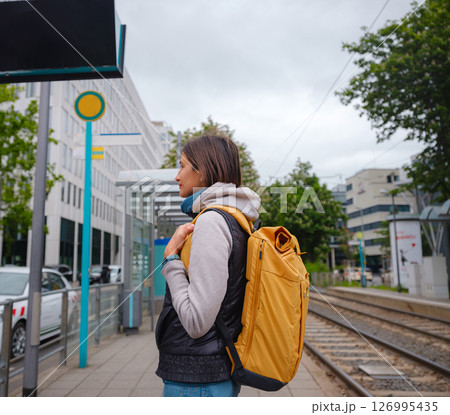 Tourist woman stands at tram stop in Frankfurt, waiting for public transportation amidst urban setting, surrounded by city life and modern buildings, experiencing everyday rhythm of city. 126995435