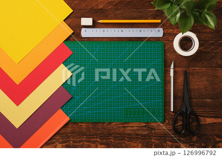 Top view of table for paper crafts. Crafting tools. Neatly arranged sheets of colored paper, green cutting mat, scissors, craft knife, pencil, and ruler. Wooden background, space for your text. Top view of table for paper crafts. Crafting tools. Neatly arranged sheets of colored paper, green cutting mat, scissors, craft knife, pencil, and ruler. Wooden background, space for your text. 126996792