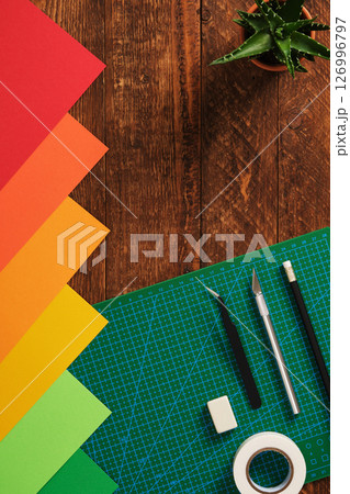 Paper crafts. Multicolored sheets of paper, green cutting mat, scissors, craft knife, pencil, glue, ruler. Wooden background, space for your text. Paper crafts. Multicolored sheets of paper, green cutting mat, scissors, craft knife, pencil, glue, ruler. Wooden background, space for your text. 126996797
